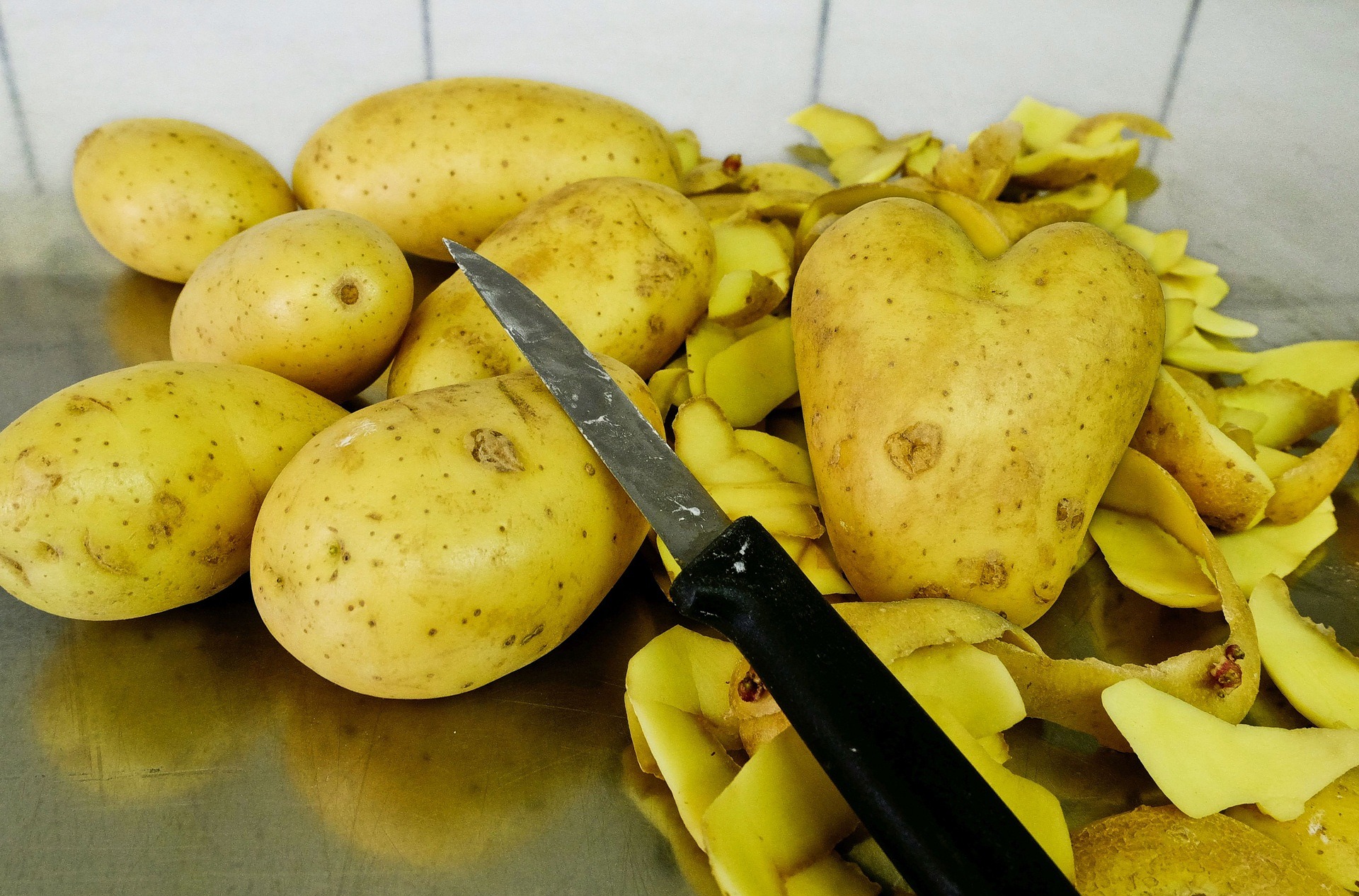 A paring knife, a bunch of potatoes, and potato peelings on a stainless steel table.